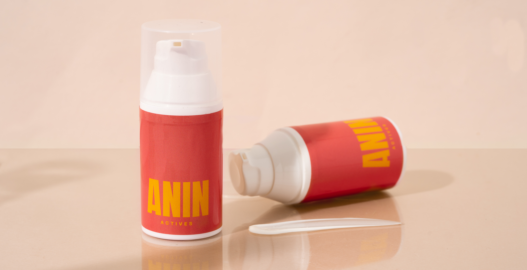 Two ANIN Actives personalized prescription bottles one standing upright the other to the left placed on the side. A white sample of the ANIN Actives prescription cream is spread in front on the ANIN Actives bottle. 