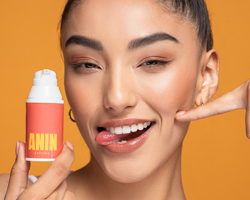 Asian model in the center against a yellow background holding a ANIN Actives personalized prescription bottle in her right hand. The ANIN Actives personalized prescription bottle has a white dispenser and a red label with a yellow ANIN Actives logo.